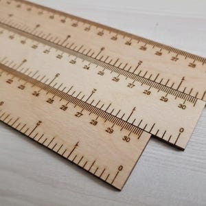 Può includere: Tre righelli di legno, ciascuno fino a 76 cm, sono esposti ad angolo. I righelli sono in legno chiaro e presentano segni neri per centimetri e millimetri. I righelli servono per misurare.