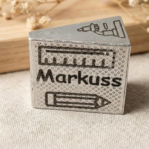 May include: A silver-colored, cube-shaped stamp with the name "Markuss" in bold black letters. The stamp features etched illustrations of a ruler, pencil, and a tool. The stamp is resting on a beige fabric surface.