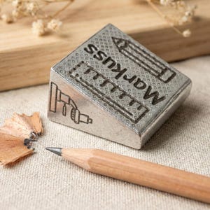 May include: A silver metal pencil sharpener with the word "Markuss" and pencil illustrations. A sharpened pencil and wood shavings are next to the sharpener. The sharpener is angled on a beige surface.