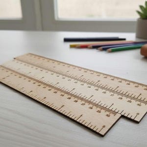 Könnte beinhalten: Drei Holzlineale, jeweils bis zu 76 cm lang, liegen auf einer hellen Oberfläche. Die Lineale sind aus hellem Holz mit dunklen Markierungen gefertigt. Buntstifte und ein kleines Notizbuch befinden sich im Hintergrund.