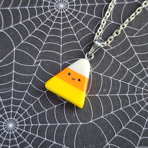May include: A silver-colored necklace with a pendant shaped like a candy corn. The pendant has a smiling face and is colored yellow, orange, and white. The background is a black surface with a white spiderweb design.