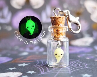 May include: A small glass bottle with a cork top and a silver clasp. Inside the bottle is a small, yellow, glow-in-the-dark ghost with a happy face. The bottle is sitting on a black and white patterned surface. "Glow in the dark!" is written on a circle around the bottle.