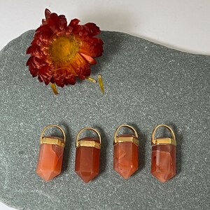 Peut inclure: Quatre pendentifs en cristal de cornaline avec des embouts dorés. Les pendentifs sont en forme de pointe et sont disposés en rangée sur une surface de pierre grise.