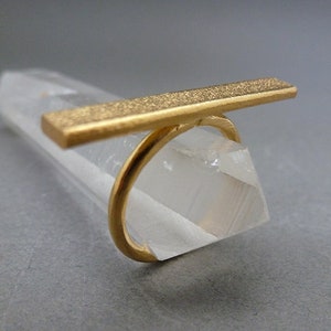 May include: A gold ring featuring a rectangular bar design. The ring's band curves around, supporting the textured bar that extends horizontally. The ring is displayed on a clear, crystalline structure, set against a neutral background.