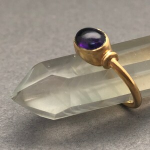 May include: A gold ring with a purple gemstone set in a bezel setting. The ring is on a clear crystal point.