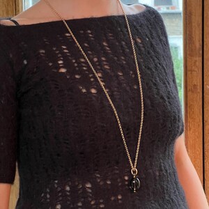 May include: A long gold-tone necklace with a dark, oval-shaped pendant. The necklace hangs over a black, open-knit sweater. The pendant appears to be a dark gemstone or glass. The chain is delicate and the overall style is elegant.