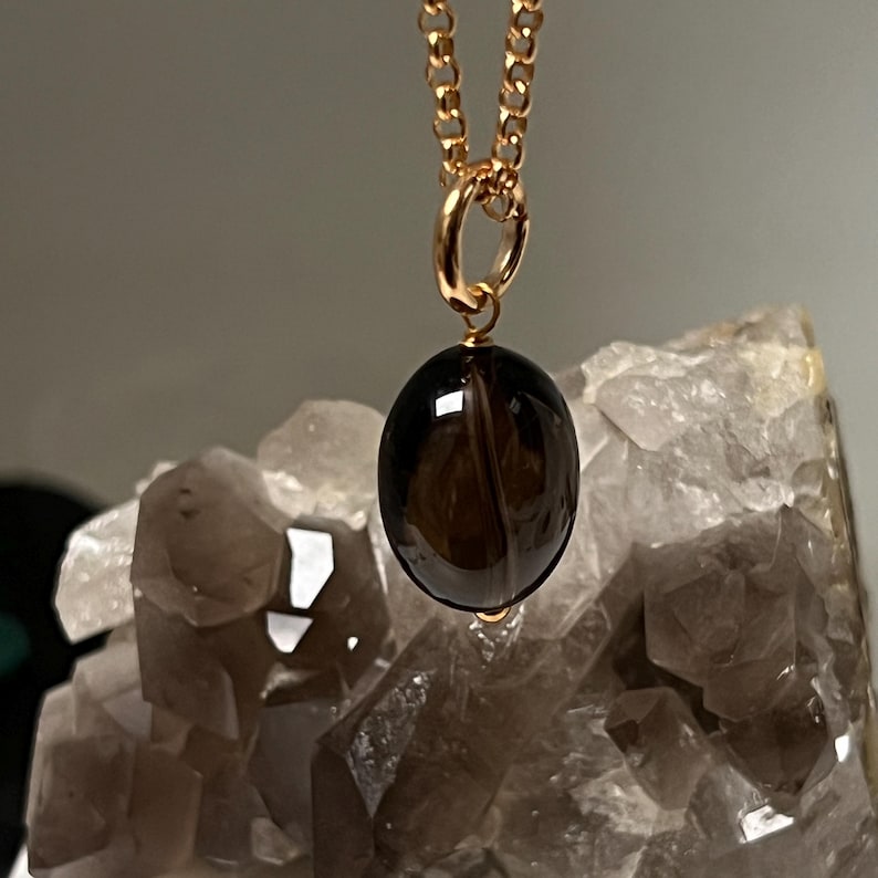 May include: A gold-colored chain necklace with a dark brown, oval-shaped pendant. The pendant is smooth and polished, with a subtle, translucent quality. The necklace is displayed against a backdrop of light-colored, crystalline formations.