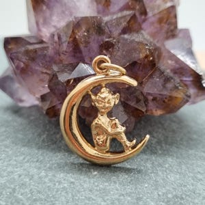 Puede incluir: Colgante dorado con una figura de elfo sentado en una luna creciente. El elfo tiene orejas puntiagudas y piernas cruzadas. El colgante está suspendido de un pequeño lazo. El fondo presenta una formación de cristal púrpura.