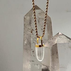 Puede incluir: Un collar de cadena de cuerda en tono dorado con un colgante transparente en forma de bala y una tapa de metal dorado. El colgante cuelga de una anilla dorada que se conecta a la cadena de metal trenzado, creando una pieza de joyería sencilla.