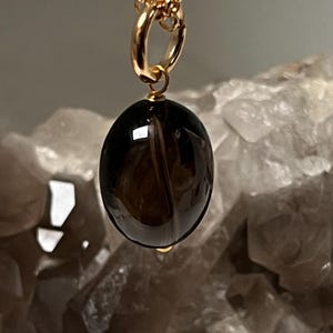 May include: A gold-colored chain necklace with a pendant. The pendant is a polished, oval-shaped, dark brown gemstone. The necklace is displayed against a blurred background of a light-colored, crystalline structure.