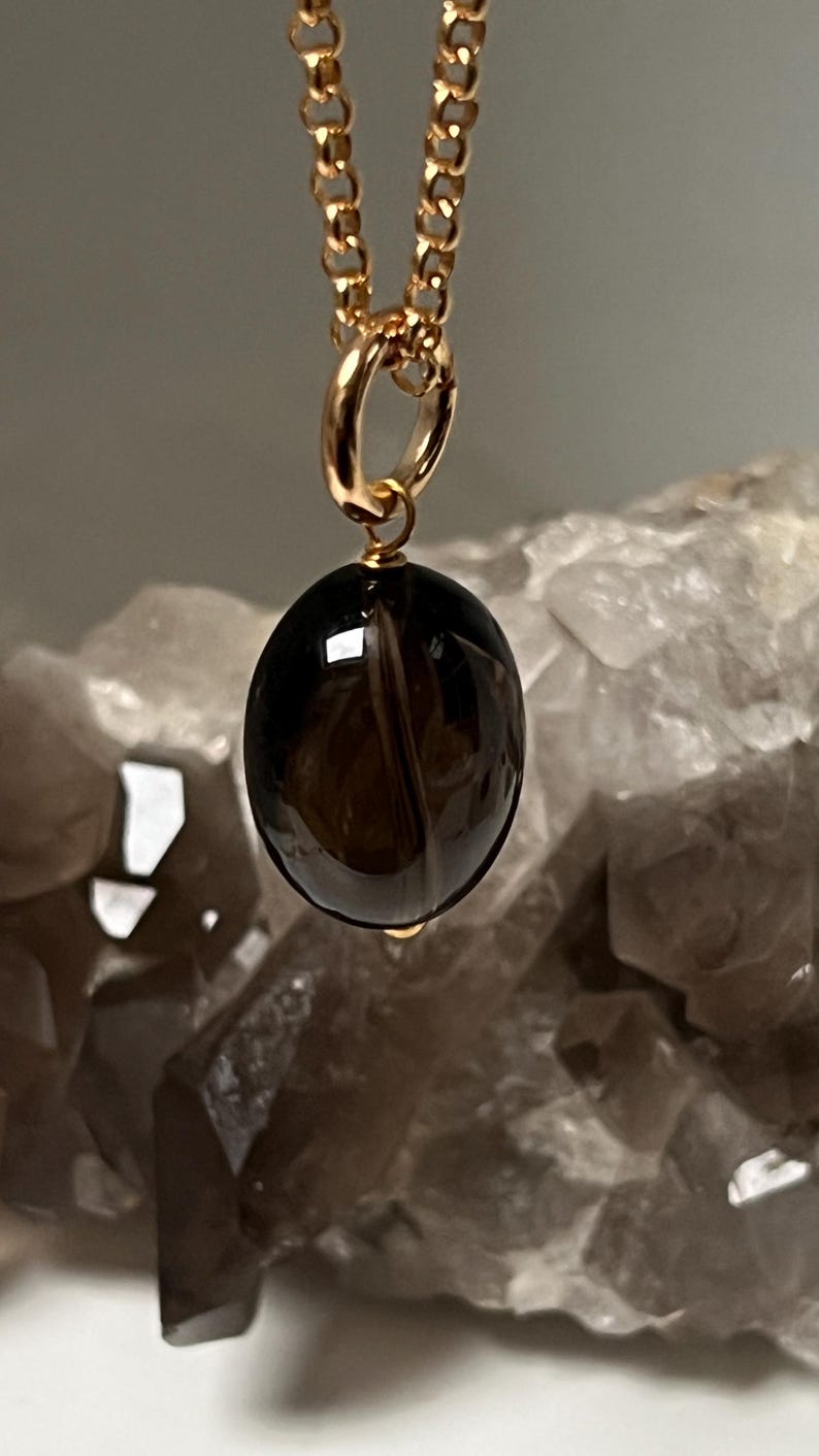 May include: A gold-colored chain necklace with a pendant. The pendant is a polished, oval-shaped, dark brown gemstone. The necklace is displayed against a blurred background of a light-colored, crystalline structure.