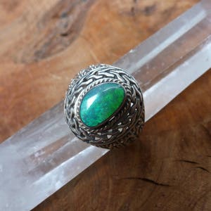 May include: A silver ring featuring a green opal gemstone set within an elaborate, intricate design.