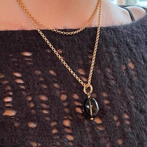 May include: A gold-colored necklace with two chains. The pendant is an oval-shaped, dark-colored stone. The necklace is worn against a dark, textured background.