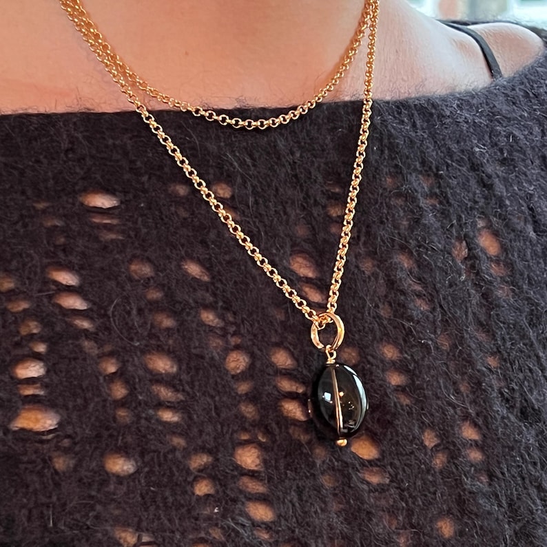 May include: A gold-colored necklace with two chains. The pendant is an oval-shaped, dark-colored stone. The necklace is worn against a dark, textured background.