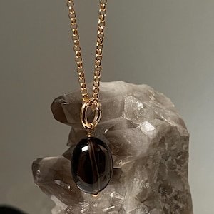 May include: A gold-colored chain necklace with a dark brown, oval-shaped pendant. The pendant is suspended from a gold-colored bail. The background is a light gray, textured surface. The necklace is a piece of jewelry.