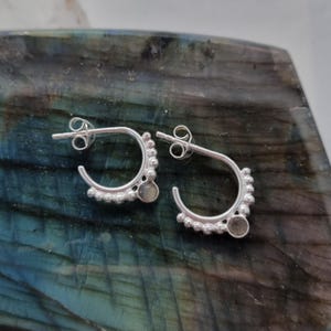 Peut inclure: Une paire de boucles d'oreilles créoles en argent. Chaque boucle d'oreille a un design incurvé avec de petites perles rondes le long du bord inférieur et une petite pierre ronde. Les boucles d'oreilles sont présentées sur une surface marbrée bleue et verte.