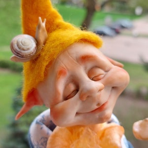 May include: A whimsical felt gnome doll with a yellow hat, closed eyes, and a smiling expression. The gnome is wearing a blue jacket with a white collar and a fluffy orange scarf. The gnome is holding a small mushroom in its hand.