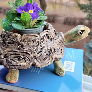 Könnte beinhalten: Ein Keramik-Schildkröten-Pflanztopf mit einem strukturierten Schalenmuster, der eine kleine Topfpflanze mit lila Blüten hält. Der Topf steht auf einem blauen Buch.