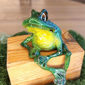 Peut inclure: Une figurine de grenouille en verre brillant avec un dos vert vif et une gorge jaune éclatant. La grenouille possède de grands yeux expressifs et est représentée en position assise, ses longues pattes fines pendant sur le bord d'un bloc en bois.