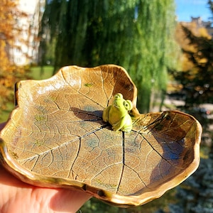 Puede incluir: Un plato de cerámica con forma de hoja con una figurita de rana verde en la parte superior. El plato tiene un esmalte marrón y verde con un patrón de hoja detallado.