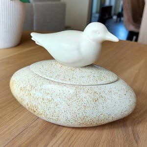 Puede incluir: Un recipiente de cerámica con tapa y base ovalada que presenta un esmalte de color crema con motas marrones. Una figura de pájaro blanca, lisa y sólida, está fijada en la parte superior de la tapa y sirve como asa para este recipiente decorativo.