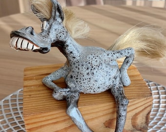 May include: A whimsical ceramic donkey figurine with a speckled gray body, a blonde mane and tail, and a wide, toothy grin. The donkey is mounted on a small, rectangular wooden block. The figurine has a cartoonish design and is posed in a playful stance.