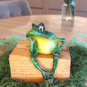 Może przedstawiać: Błyszcząca ceramiczna figurka żaby z żywo zielonym grzbietem i jasnym żółtym brzuchem. Żaba przedstawiona jest w pozycji siedzącej, z jedną ręką przy brodzie, a drugą na biodrze, z dużymi oczami i szczegółowo odwzorowanymi błonami pławnymi.