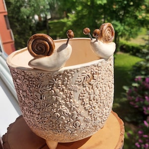 Könnte beinhalten: Cremefarbener Keramik-Blumentopf mit geprägtem Blumenmuster und drei kleinen Füßen. Zwei Schneckenfiguren mit braunen Gehäusen und Fühlern sitzen am Rand. Der Topf steht auf einer Holzscheibe.