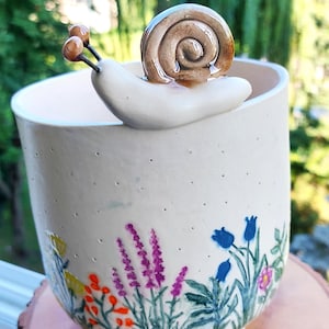 Può includere: Vaso in ceramica con una figura di lumaca. Il vaso bianco sporco presenta un motivo floreale dipinto a mano in colori vivaci. La lumaca ha un guscio marrone e si trova sul bordo, aggiungendo un tocco di fascino ispirato alla natura.