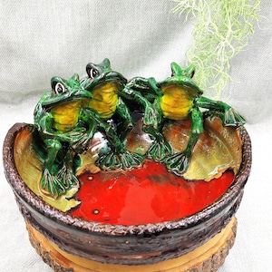 May include: Three green ceramic frogs sitting on the edge of a red and brown ceramic bowl. The bowl is on a wooden base with a bark texture.