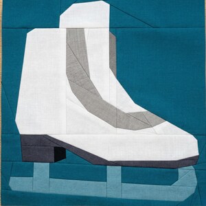 May include: A fabric art piece featuring a white and gray ice skate design on a teal background. The skate is detailed with geometric shapes and lines, set against a solid teal square. The artwork is likely a quilt block or a similar textile art.