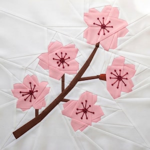 Puede incluir: Un bloque de edredón cuadrado de tela con un diseño de papel ensamblado que muestra una rama marrón con cuatro flores de cerezo rosas. Cada flor tiene cinco pétalos y un centro bordado en rojo oscuro sobre un fondo blanco.