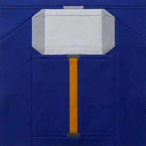 May include: A blue fabric square featuring a stylized hammer design. The hammer head is light gray, with darker gray accents. The handle is a vertical orange stripe, with gray details at the top and bottom. The background is a solid, deep blue.