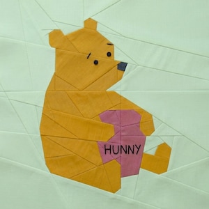 Op de afbeelding: Een patchworkontwerp met een gele beer die een roze honingpot vasthoudt met het woord "HUNNY" in zwart. De beer staat tegen een lichtgroene achtergrond met geometrische lijnen.