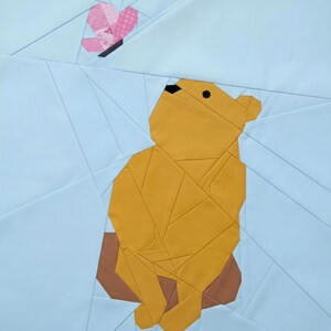 May include: A patchwork quilt square featuring a geometric design of a yellow bear sitting and looking up at a pink butterfly. The background is light blue with geometric lines.