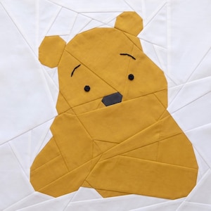 May include: A geometric quilt block featuring a yellow bear with black button eyes and a black nose. The bear is set against a white background with geometric lines. The bear's body is made of various yellow fabric shapes.