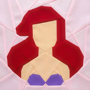 May include: A fabric block featuring a geometric design of a character with red hair, a tan face and neck, and a purple top. The character is set against a pink and white background, with a green cutting mat visible.