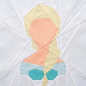 May include: A fabric quilt block featuring a geometric design of a woman's face. The face is peach-colored, with blonde hair and a turquoise bodice. The background is white, with angular lines radiating outwards.
