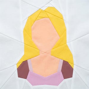 May include: A fabric quilt block featuring a geometric design of a person's face. The hair is yellow, the face is peach, and the clothing is pink and brown. The background is white, and the block is square.