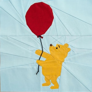 Könnte beinhalten: Ein Patchwork-Quilt-Quadrat mit einem gelben Bären, der einen roten Ballon vor einem hellblauen Hintergrund hält. Der Bär und der Ballon bestehen aus geometrischen Formen, mit einer schwarzen Schnur, die den Ballon bindet. Der Hintergrund hat ein strahlenförmiges Design.