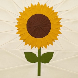 May include: A fabric block featuring a detailed sunflower design. The sunflower has a brown center, yellow petals, green stem, and leaves. The background is a light cream color, with geometric lines radiating from the flower.