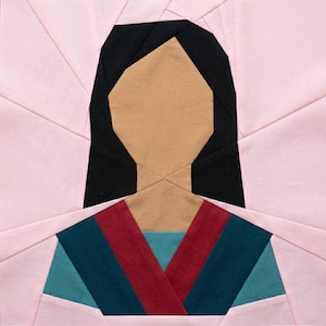 May include: A fabric block featuring a geometric design of a person's face and upper body. The face is tan with black hair. The clothing is teal, red, and blue. The background is light pink.