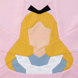 May include: A fabric block featuring a geometric design of a character's face. The design includes yellow hair, a black headband, a light peach face, and a blue and white dress. The background is light pink.