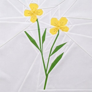 May include: A fabric quilt block featuring two yellow flowers with green stems and leaves against a white background. The flowers have detailed yellow centers. The white background is composed of geometric shapes.