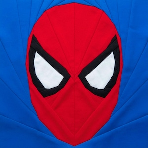 May include: A fabric quilt block featuring the face of a superhero. The design includes a red mask with white eyes outlined in black, set against a blue background. The quilt block is square and appears to be handmade.