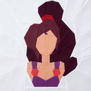 May include: A geometric fabric art piece featuring a stylized portrait of a woman. The design uses various shades of purple, red, and peach against a white background. The woman's hair is dark purple, and she wears a purple top with red accents.
