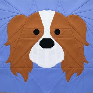 May include: A square fabric block featuring a geometric design of a Cavalier King Charles Spaniel dog's face. The dog's face is brown, white, and black, set against a light blue background. The design is created with various fabric shapes.