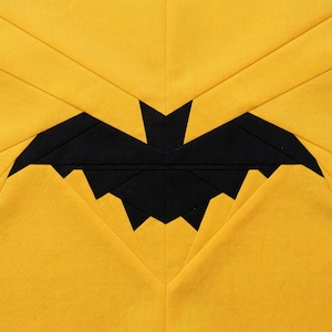 May include: A Halloween-themed fabric block featuring a black bat silhouette against a yellow background. The bat is centered, with the yellow fabric folded to create a geometric design. The block is square.