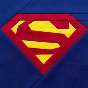 May include: A fabric block featuring the Superman logo. The design includes a red diamond shape with a yellow 'S' emblem, set against a blue background. The block is made of fabric and is likely part of a quilt or craft project.