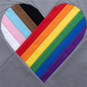 May include: A square fabric quilt block featuring a central heart shape constructed from diagonal strips of fabric. The heart displays the Progress Pride flag colors on the left and the traditional rainbow flag colors on the right, set against a gray background.